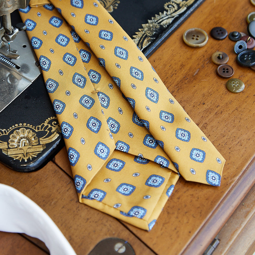 
                  
                    Cravatta fantasia in pura seta senape con disegno azzurro e bianco
                  
                